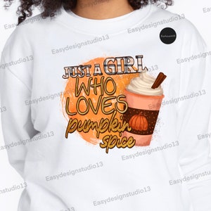 Pode incluir: Camisola branca com um design gráfico que diz "Just a girl who loves pumpkin spice" com uma ilustração estilizada de um latte de especiarias de abóbora.