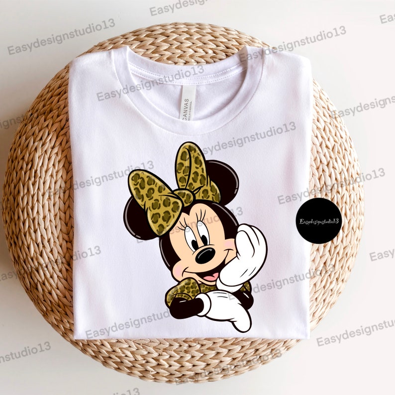 May include: A white t-shirt with a cartoon image of Minnie Mouse wearing a leopard print bow. Minnie Mouse is smiling and has her hands up in a gesture of greeting.