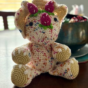 May include: A crocheted triceratops toy made from cream-colored yarn with multicolored speckles. It features three dark pink strawberry-shaped horns on its head, green leaf accents, black safety eyes, and tan-colored feet with white claws.