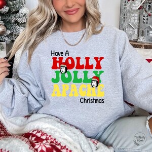 May include: A light gray sweatshirt with the text "Have A HOLLY JOLLY APACHE Christmas" in red, green, and yellow. The design includes illustrations of a person wearing a Santa hat. The sweatshirt is a festive holiday apparel item.