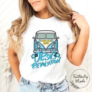 May include: White t-shirt with a light blue VW bus graphic. The design includes the text "LOVE LIKE JESUS" and "JESUS REVOLUTION", along with blue and yellow flowers. A casual wear item.
