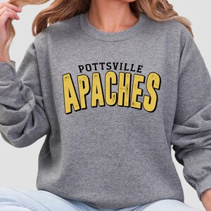May include: A gray sweatshirt with the text "POTTSVILLE APACHES" in black and yellow block letters. The sweatshirt is a crew neck style and appears to be made of a soft material. The design is centered on the chest.