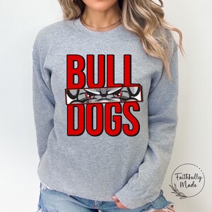 May include: Gray sweatshirt with the words "BULLDOGS" in red letters. The word "BULL" is above the word "DOGS" and a bulldog's face is partially visible between the words.