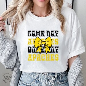 May include: White t-shirt with the text "GAME DAY APACHES" in black and yellow, featuring a yellow bow graphic. The shirt is a classic crew neck style.