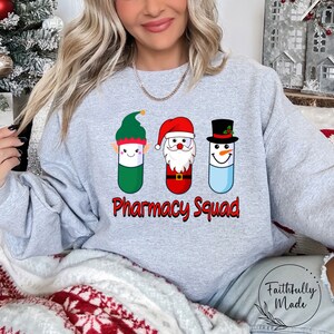 Op de afbeelding: Een grijs sweatshirt met een kerstthema ontwerp met een elf, de kerstman en een sneeuwpop, allemaal afgebeeld als pillen. De tekst "Pharmacy Squad" is onder de afbeeldingen gedrukt.