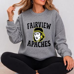 Könnte beinhalten: Ein graumeliertes Sweatshirt mit Rundhalsausschnitt, schwarzem Schriftzug FAIRVIEW APACHES und einer gelb-schwarzen Grafik einer Person mit einem Stirnband, auf dem das Wort APACHES steht, kombiniert mit einer schwarzen Leggings und weißen Turnschuhen.