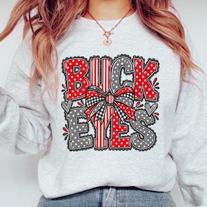 May include: A light gray crewneck sweatshirt featuring the text BUCKEYES in a bold, block font. The letters alternate between red with white stripes and gray with white polka dots, all outlined in black. A plaid bow sits in the center.