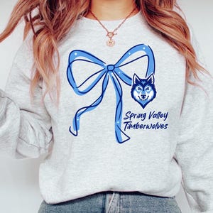 May include: A light gray crewneck sweatshirt featuring a large blue graphic of a ribbon bow, a wolf head illustration, and the text Spring Valley Timberwolves printed in blue. The design includes small white star accents within the blue bow.