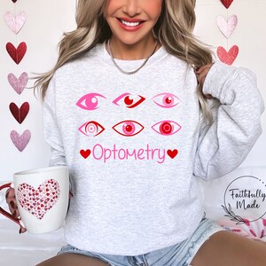 May include: A gray sweatshirt with a pink and red eye design and the text "Optometry" with hearts on either side. The sweatshirt is being worn by a person.