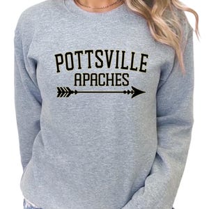 Puede incluir: Sudadera gris claro con las palabras "POTTSVILLE APACHES" en letras negras sobre un gráfico de flecha negra. La sudadera tiene cuello redondo y mangas largas.