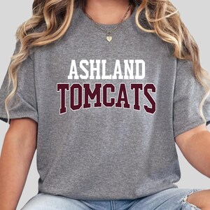 Może przedstawiać: Szara koszulka z napisem "ASHLAND" białymi literami nad napisem "TOMCATS" bordowymi literami. Koszulka ma okrągły dekolt.