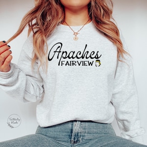 Może przedstawiać: Szara bluza z napisem "Apaches Fairview" czarnymi literami. Po prawej stronie napisu znajduje się mały obrazek osoby w opasce na głowie.