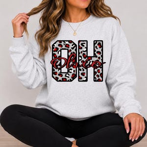 May include: A light gray crewneck sweatshirt featuring the block letters OH in a gray, black, and red leopard print pattern. The word Ohio is written in a red cursive script font across the center of the letters.
