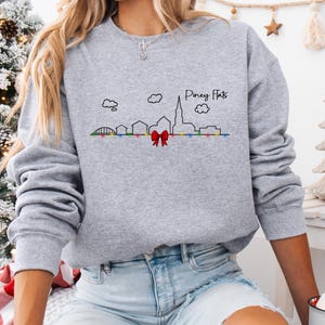 May include: A heather gray sweatshirt featuring a skyline design with the text "Piney Flats." The design includes a red bow, colorful lights, and black outlines of buildings and clouds. The sweatshirt is a long-sleeved, crew neck style.