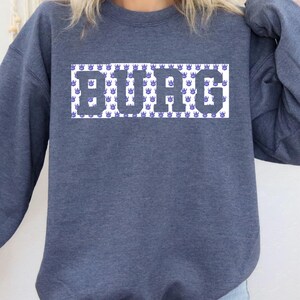 May include: A heathered navy blue crewneck sweatshirt featuring a rectangular graphic on the chest. The graphic displays the word BURG in large, dark grey block letters set against a white background patterned with small, repeating purple Viking helmet icons.