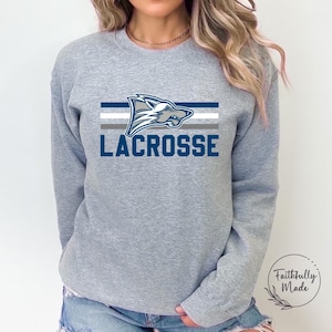 Könnte beinhalten: Grauer Sweatshirt mit einem blauen Wolfskopf-Motiv und dem Wort "LACROSSE" in blauen Buchstaben. Der Wolfskopf ist von fünf horizontalen Streifen in Weiß, Grau und Blau umgeben.