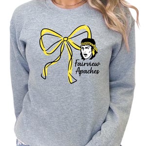 Puede incluir: Sudadera gris con un lazo amarillo y el texto "Fairview Apaches". El diseño incluye un gráfico de una persona con una cinta en la cabeza. La sudadera es de cuello redondo. Prenda casual.