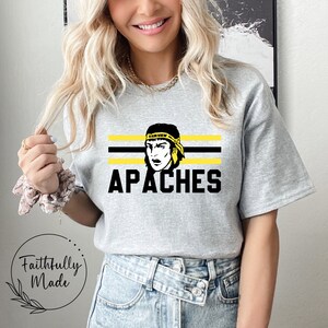 Puede incluir: Camiseta gris con rayas negras y amarillas y una ilustración en blanco y negro de una persona con una diadema que dice "Fairview" y la palabra "APACHES" en letras negras.