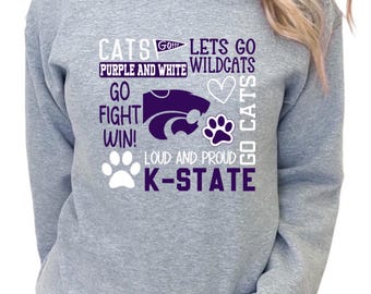 以下が含まれることがあります： 白と紫で「CATS GO WILDCATS」と「K-STATE」の文字が入ったヘザーグレーのスウェットシャツ。デザインには足跡、ハート、「GO FIGHT WIN！」の文字が含まれています。