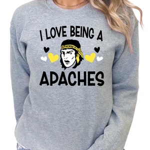 May include: A light gray sweatshirt with the text "I LOVE BEING A APACHES" in black. The design includes a graphic of a person with a yellow headband, black hair, and black and yellow hearts.