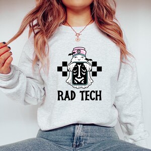 May include: Light gray sweatshirt with a graphic featuring a cartoon ghost in a medical cap and a skeleton design. The text "RAD TECH" is printed below the graphic in black. The design includes black and white checkered squares.