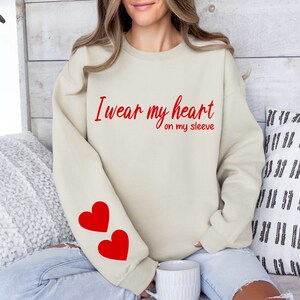 Może przedstawiać: Beżowa bluza z napisem "I wear my heart on my sleeve" w czerwonych literach. Dwa czerwone serca są nadrukowane na lewym rękawie.