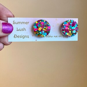 May include: A pair of round stud earrings with a colorful confetti design. The earrings feature a mix of pink, teal, gold, and iridescent glitter. The earrings are displayed on a white card with the text "Summer Lush Designs".
