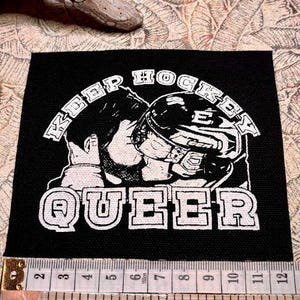 Può includere: Toppa quadrata nera con testo e grafica bianchi. Il testo recita "KEEP HOCKEY QUEER" in un font stilizzato. La grafica raffigura due figure con l'attrezzatura da hockey. La toppa è larga circa 7,6 cm.