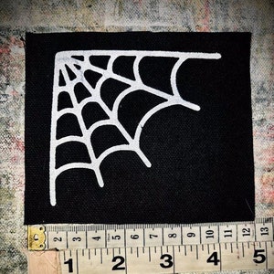 以下が含まれることがあります： 角に白いクモの巣のデザインが施された黒い布のパッチ。パッチの幅は約10cmです。
