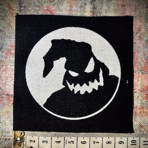 May include: Black and white iron-on patch featuring a silhouette of the character Oogie Boogie from the movie The Nightmare Before Christmas. The character is depicted with a large grin and a pointed hat.