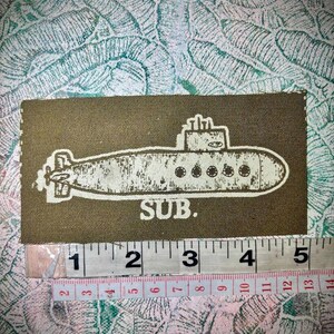 May include: A rectangular olive green fabric patch featuring a white screen-printed illustration of a submarine and the text "SUB." positioned above a measuring tape showing a width of approximately 5 inches and 13 centimetres.