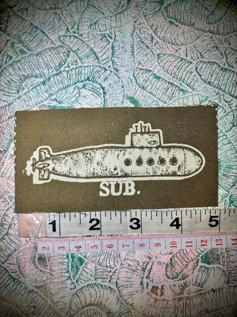 May include: A rectangular olive green fabric patch featuring a white screen-printed illustration of a submarine and the text "SUB." positioned above a measuring tape showing a width of approximately 5 inches and 13 centimetres.