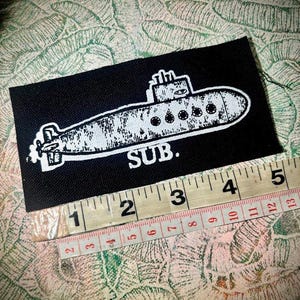 May include: A rectangular black fabric patch featuring a white screen-printed illustration of a submarine with the text "SUB." below it. A measuring tape is positioned along the bottom edge, showing the patch is approximately 11.5 cm long.