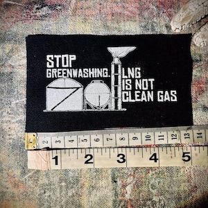 May include: Black patch with white text that reads "STOP GREENWASHING. LNG IS NOT CLEAN GAS". The patch features a white silhouette of a gas processing plant.