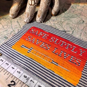 May include: Embroidered patch with the text "SAFE SUPPLY SAVES LIVES" and a syringe design. The patch features a gradient of orange and yellow, set against a striped blue and white background. A measuring tape is in the foreground.