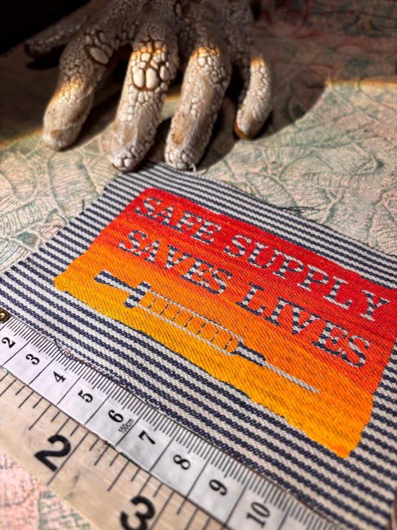 May include: Embroidered patch with the text "SAFE SUPPLY SAVES LIVES" and a syringe design. The patch features a gradient of orange and yellow, set against a striped blue and white background. A measuring tape is in the foreground.
