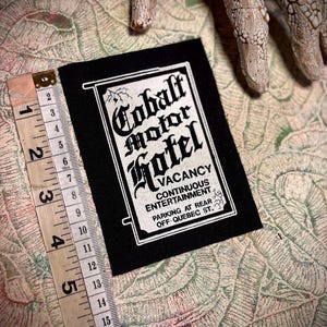 Op de afbeelding: Een zwart embleem met witte tekst met de tekst "Cobalt Motor Hotel VACANCY CONTINUOUS ENTERTAINMENT PARKING AT REAR OFF QUEBEC ST." Een meetlint toont dat het embleem ongeveer 7,6 cm lang is.