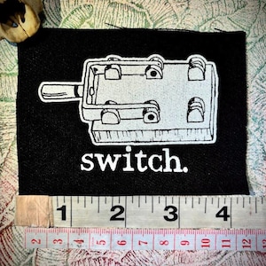 May include: Black fabric patch with a white illustration of a switch and the word "switch." The patch is approximately 4 inches wide. The design features a detailed outline of the switch mechanism.