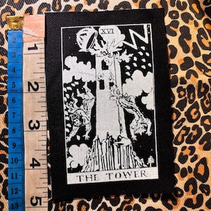 Puede incluir: Un parche rectangular de tela en blanco y negro con el diseño de la carta del tarot La Torre, el número romano XVI en la parte superior y el texto THE TOWER en la parte inferior. Una cinta métrica azul muestra que mide 15 cm de largo.
