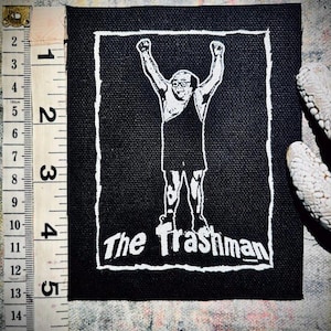 May include: Black and white iron-on patch with a cartoon image of a man with his arms raised in the air. The text "The Trashman" is printed below the image.