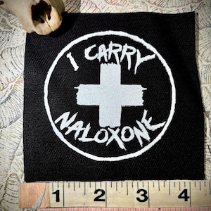 Op de afbeelding: Een vierkante zwarte stoffen patch met een witte cirkelvormige rand, een wit kruis in het midden en de tekst I CARRY NALOXONE in een versleten lettertype. Een meetlint onderaan toont een breedte van ongeveer 10 cm.