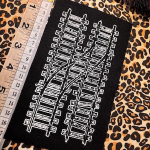 May include: Black rectangular patch with a white illustration of railway tracks. The patch is approximately 10 cm wide and 15 cm tall. The background is a leopard print fabric.