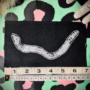 May include: Black rectangular patch with a white embroidered worm design. The patch is approximately 6 inches long. The background is a green and pink leopard print.