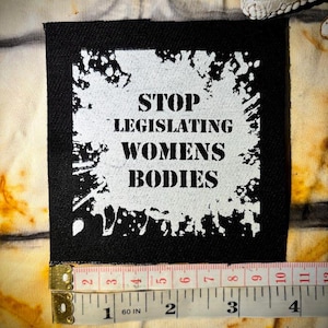 May include: Black square patch with white text that reads "STOP LEGISLATING WOMENS BODIES." The patch is approximately 4 inches wide. The background is a tie-dye pattern in shades of yellow, brown, and white.