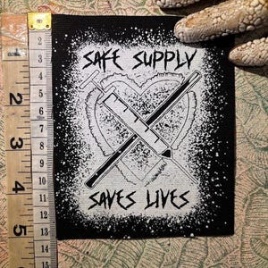 Puede incluir: Parche cuadrado negro con gráfico blanco de un corazón, dos jeringas cruzadas y el texto "SAFE SUPPLY SAVES LIVES". El parche mide aproximadamente 10 cm de ancho.