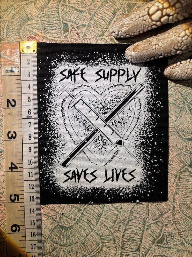 May include: Black square patch with white graphic of a heart, two crossed syringes, and the text "SAFE SUPPLY SAVES LIVES". The patch is approximately 10 cm wide.