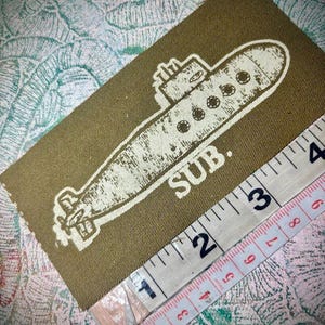 May include: A rectangular olive green fabric patch featuring a white screen-printed graphic of a submarine and the text SUB. A white measuring tape is positioned along the bottom edge, showing the patch length is approximately 10 cm.