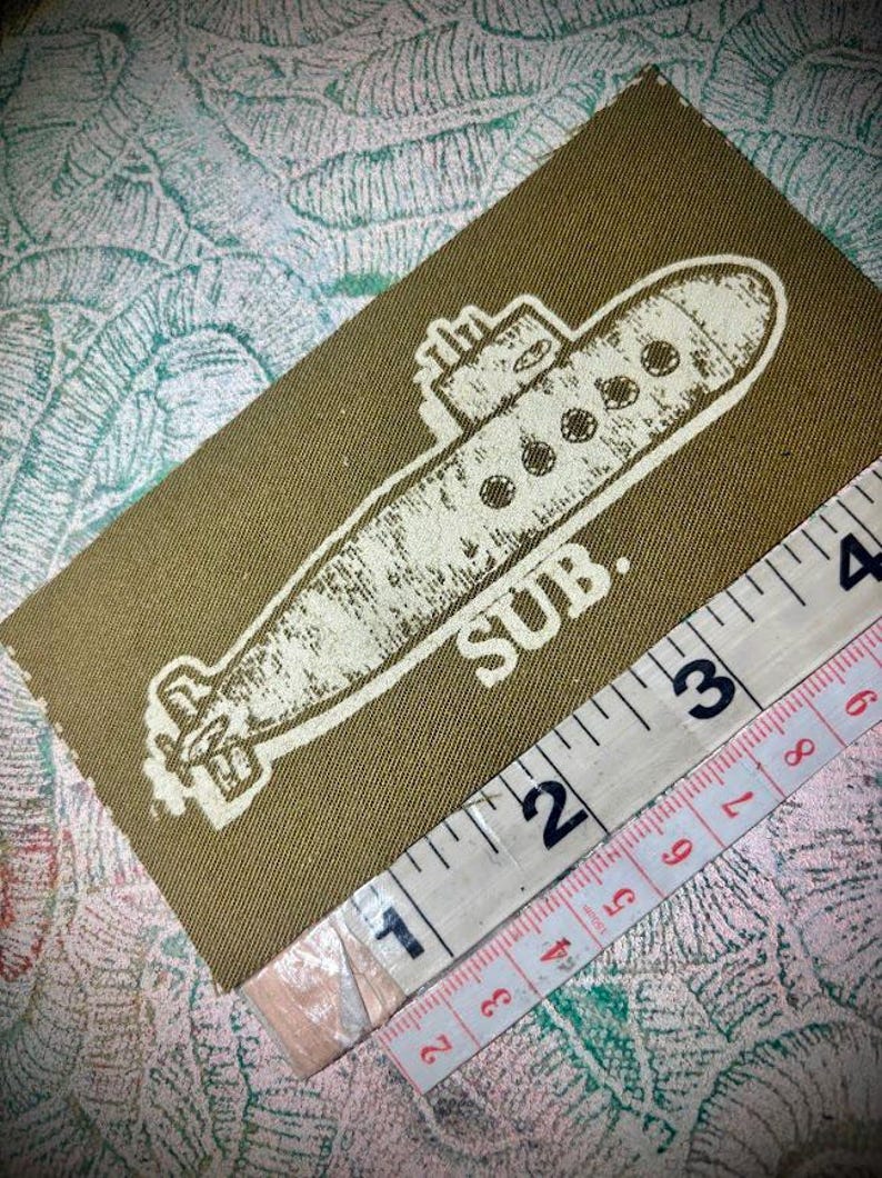 May include: A rectangular olive green fabric patch featuring a white screen-printed graphic of a submarine and the text SUB. A white measuring tape is positioned along the bottom edge, showing the patch length is approximately 10 cm.