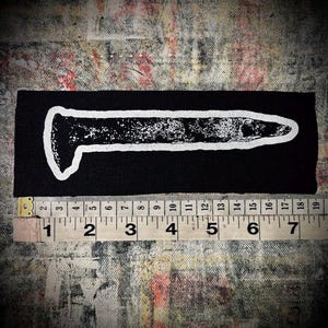 May include: A white iron-on patch with a black background featuring a stylized image of a nail. The nail is outlined in white and has a textured appearance.