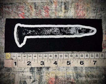 May include: A rectangular black fabric patch featuring a white screen-printed graphic of a large nail. A measuring tape is positioned along the bottom edge, indicating the patch has a total length of approximately 7.5 inches.